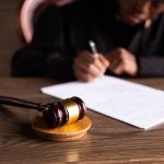 Pessoa assina documentos jurídicos sobre uma mesa, com um martelo de juiz em primeiro plano.
