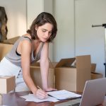 Empreendedora administra seu negócio de roupas em um escritório doméstico, diante de um laptop e caixas de produtos.
