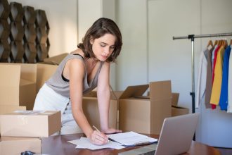 Empreendedora administra seu negócio de roupas em um escritório doméstico, diante de um laptop e caixas de produtos.