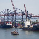 Porto industrial com grandes navios porta-contêineres e guindastes sob céu claro.